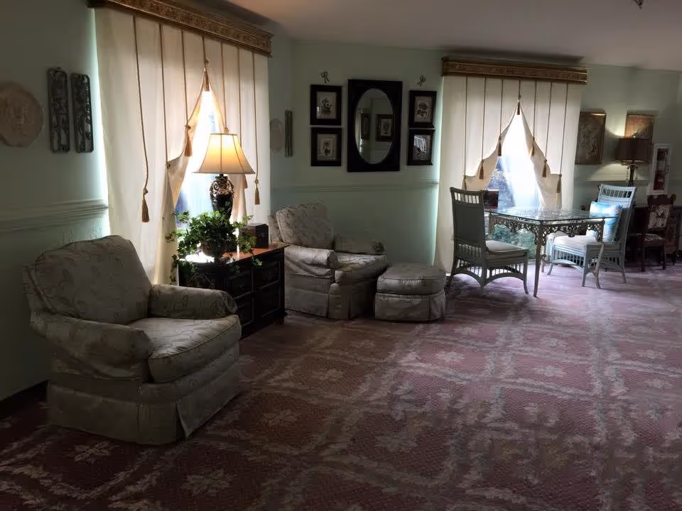 A cozy sitting area in a senior living facility with two cushioned armchairs and an ottoman, a small dark wooden side table with a decorative lamp and a potted plant, two large windows with cream-colored curtains, a glass-top table with two chairs, and framed artwork on the light green walls.