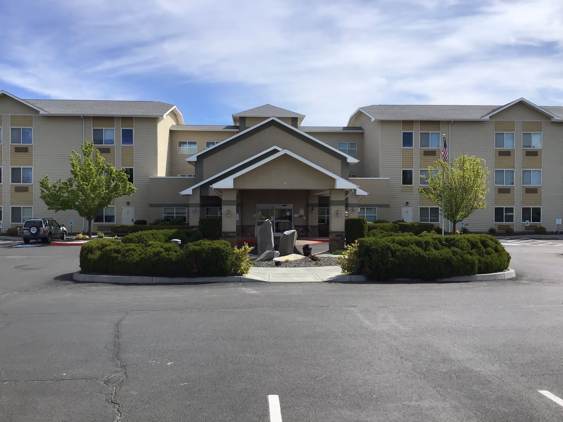 Front exterior view of a three-story senior living facility building with beige siding and a covered entrance. There are two small trees and green bushes in front, with a paved parking lot in the foreground under a partly cloudy sky.