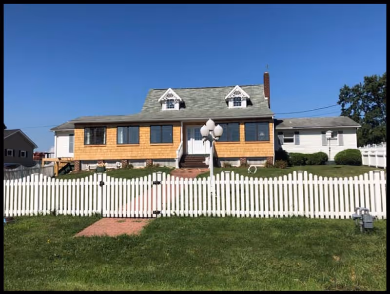 Front exterior view of a single-story house with a green shingled roof, two dormer windows, and a white picket fence in front. There is a brick pathway leading to the front door and a lamp post with three lights near the entrance. The sky is clear and blue.