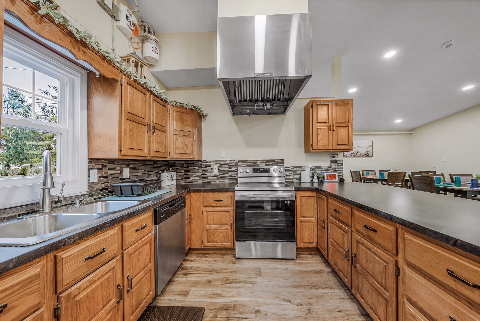 A spacious kitchen with wooden cabinets, a stainless steel stove and range hood, a dishwasher, and a double sink under a window. The kitchen has a tiled backsplash and a long countertop that extends into a dining area with tables and chairs in the background.
