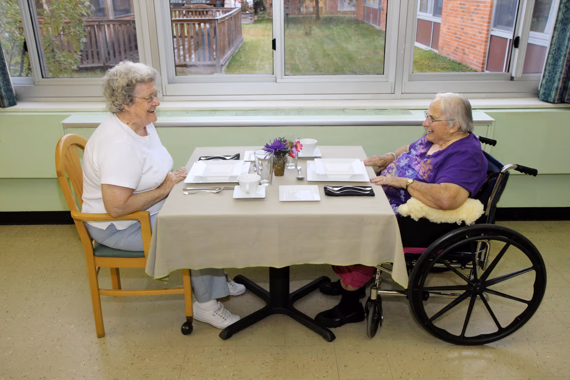 Two elderly women sitting at a table set for a meal in a dining area. One woman is seated in a wooden chair, and the other is in a wheelchair. They are smiling and engaging with each other. The table has a beige tablecloth, white plates, cups, and silverware, with a small vase of flowers in the center. Large windows behind them show an outdoor view with grass and a wooden fence.