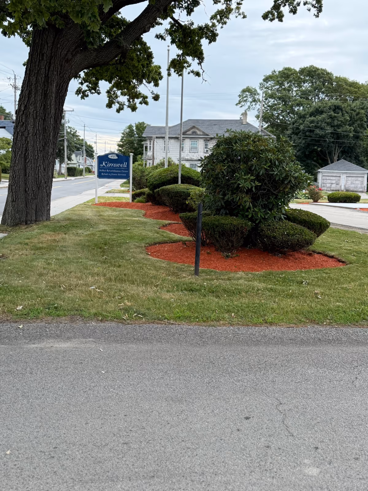 Outdoor view of the Kimwell Nursing and Rehabilitation facility sign on a grassy area with trimmed bushes and a large tree, with a building and driveway in the background under a cloudy sky.
