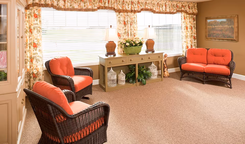 A cozy living room with three wicker chairs and a loveseat, all with orange cushions. There is a beige cabinet with drawers and decorative items including two lamps, a flower arrangement, and white birdcages. The room has floral curtains on the windows and a framed painting on the wall.