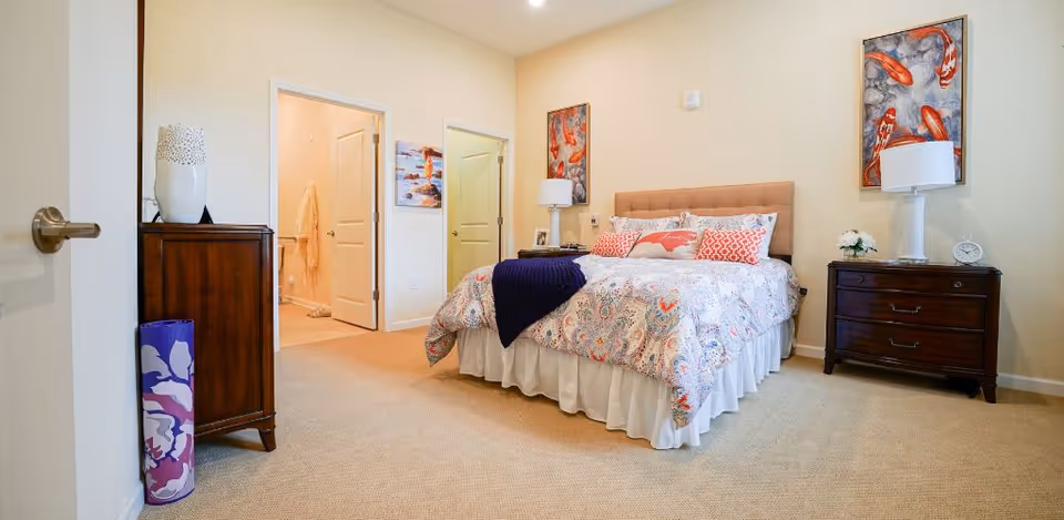 Well-lit furnished bedroom with a queen bed, two nightstands, a chest of drawers, and an open doorway to a bathroom.