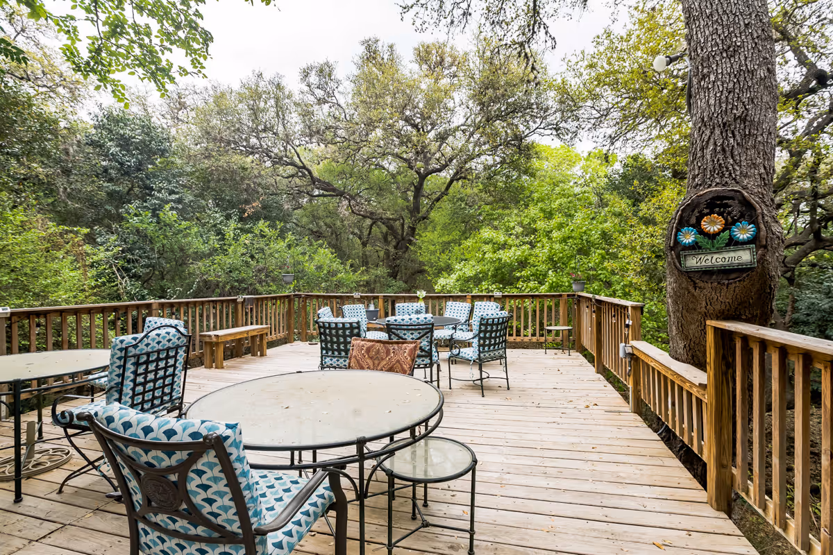 Outdoor wooden deck with multiple round glass tables and cushioned chairs featuring blue and white patterned upholstery. The deck is surrounded by wooden railings and lush green trees. A tree on the right side has a decorative sign with flowers and the word 'Welcome'.