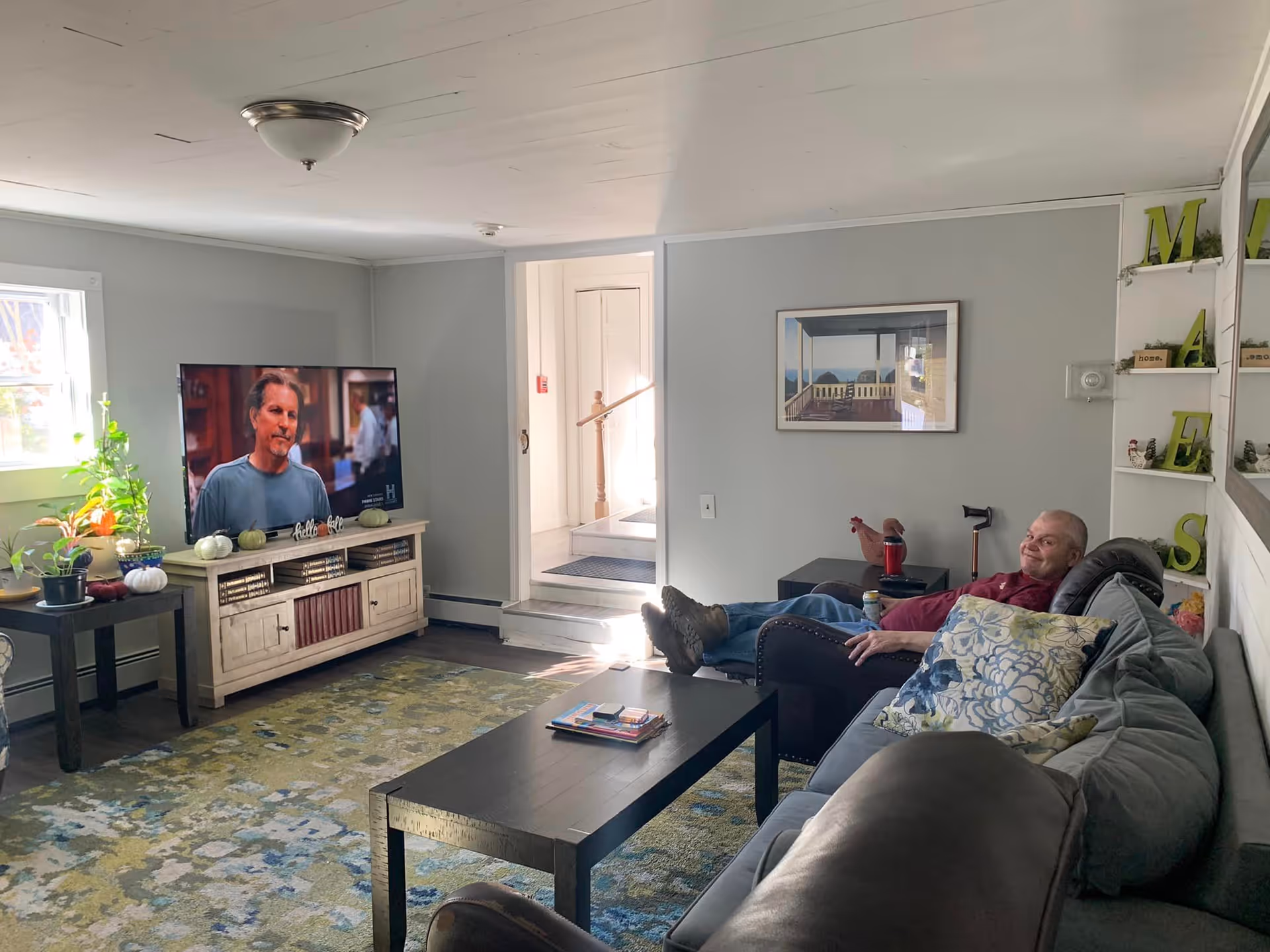 Cozy living room with a couch and recliner, coffee table, TV on a stand, potted plants, and a man reclining on the sofa.