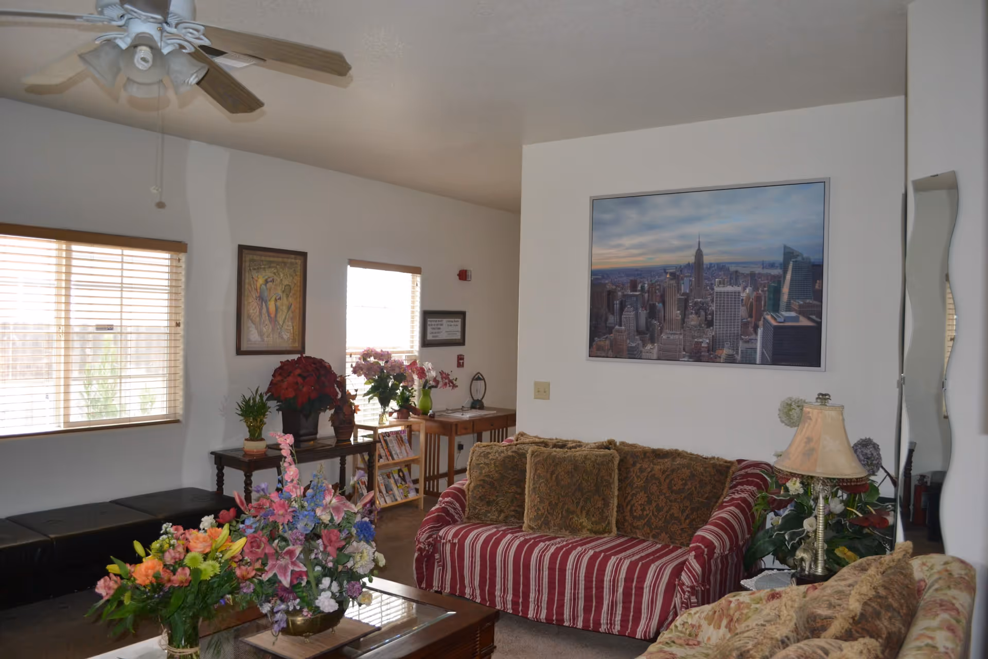 A cozy living room with patterned sofas, a coffee table covered in floral arrangements, framed wall art, and a ceiling fan.