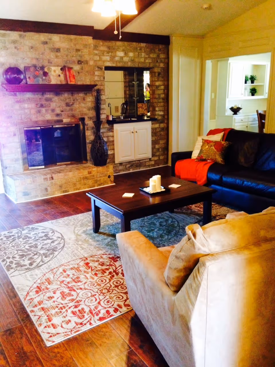 A cozy living room with a brick fireplace, a dark wooden coffee table with candles, a beige armchair, and a black leather sofa with colorful cushions and a red throw blanket. The room has wooden flooring and a patterned area rug. There is a small wet bar area with a mirror and cabinets built into the brick wall, and an open doorway leading to a dining area with white cabinetry and green plants.