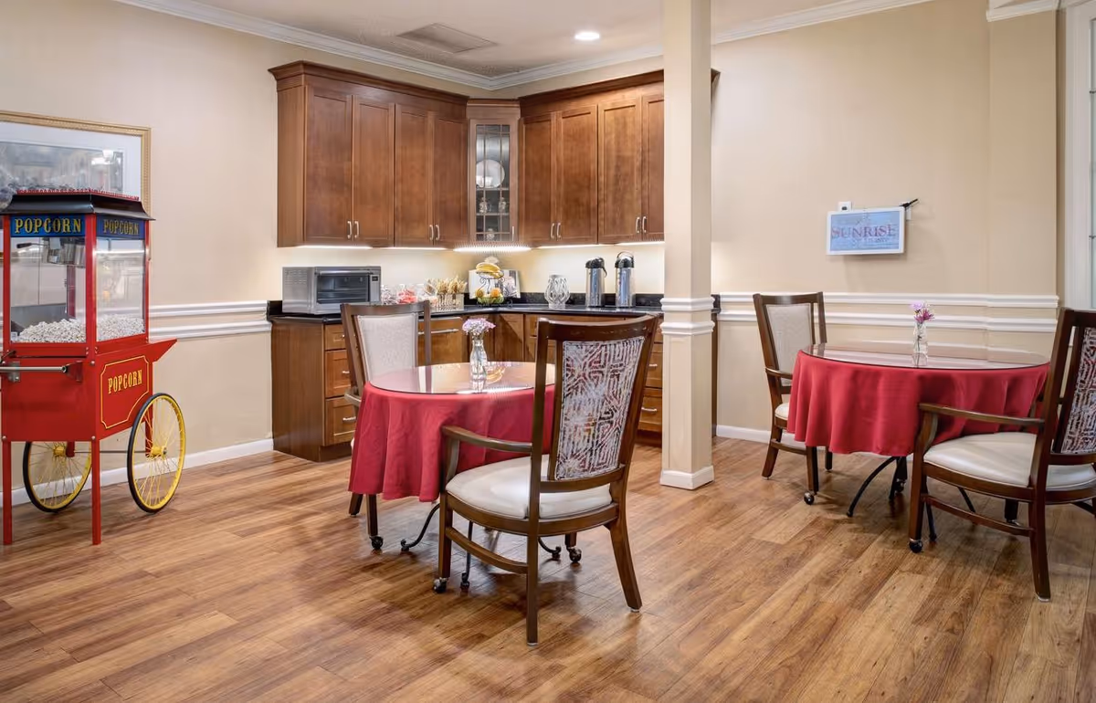 Interior view of a dining area with wooden flooring, two round tables covered with red tablecloths, each with a small vase of flowers. There are wooden chairs with cushioned seats around the tables. In the background, there is a kitchenette with wooden cabinets, a microwave, coffee dispensers, and snacks. A red popcorn machine is positioned on the left side of the image.