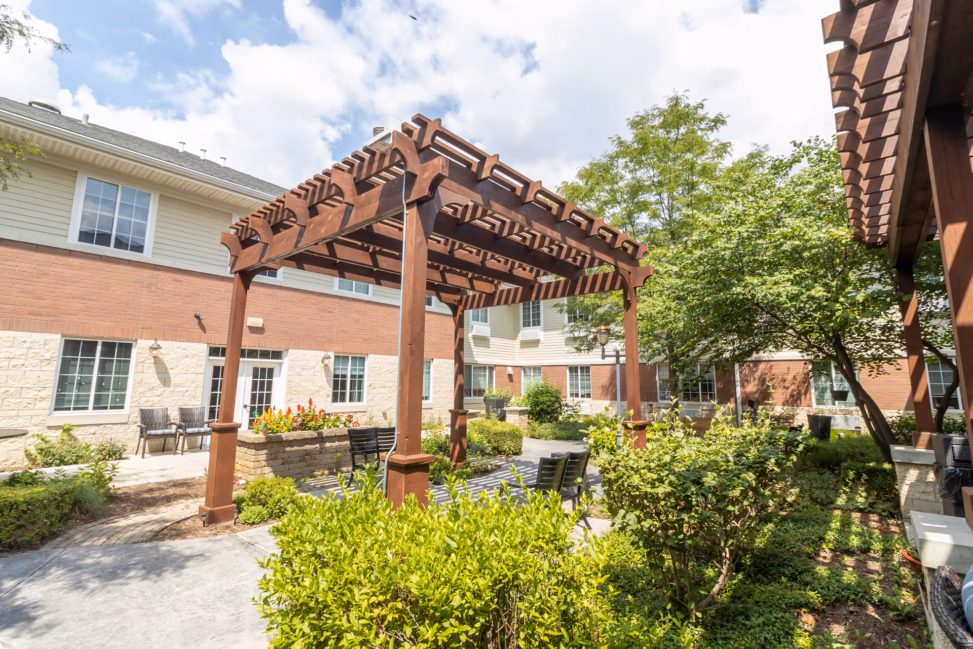 Outdoor courtyard area at a senior living facility with wooden pergolas, green shrubs, trees, and benches. The building surrounding the courtyard has brick and light-colored siding with multiple windows.
