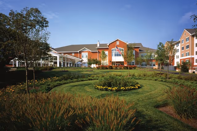 Exterior view of Ann's Choice Retirement Community building with a well-maintained garden and circular flower bed in the foreground under a clear blue sky.