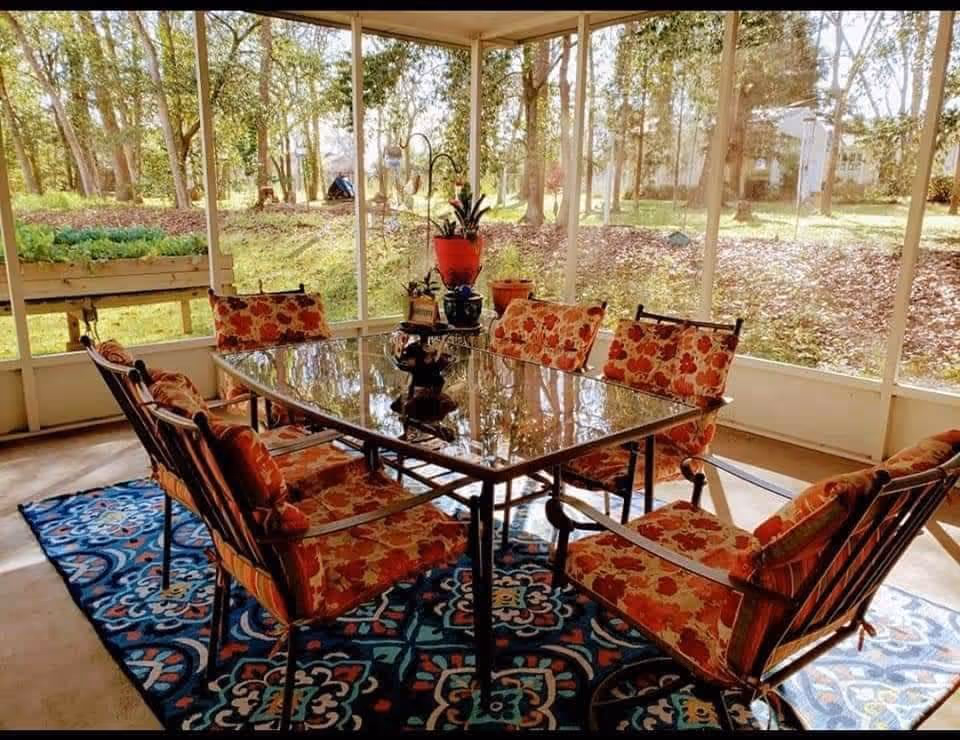 A sunlit enclosed patio with a glass-top dining table surrounded by six cushioned chairs with floral upholstery. The floor features a colorful patterned rug, and several potted plants are placed on and around the table. Large windows provide a view of trees and greenery outside.