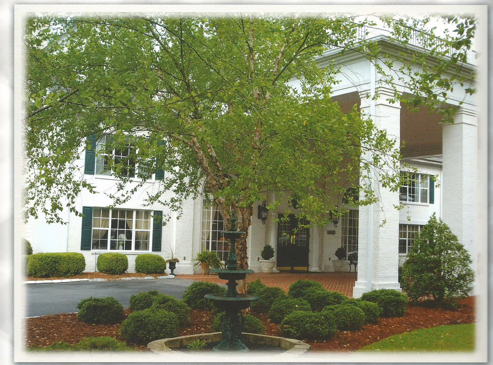 Front entrance of a white colonial-style building with large columns, a fountain, a tree, and landscaped shrubs.
