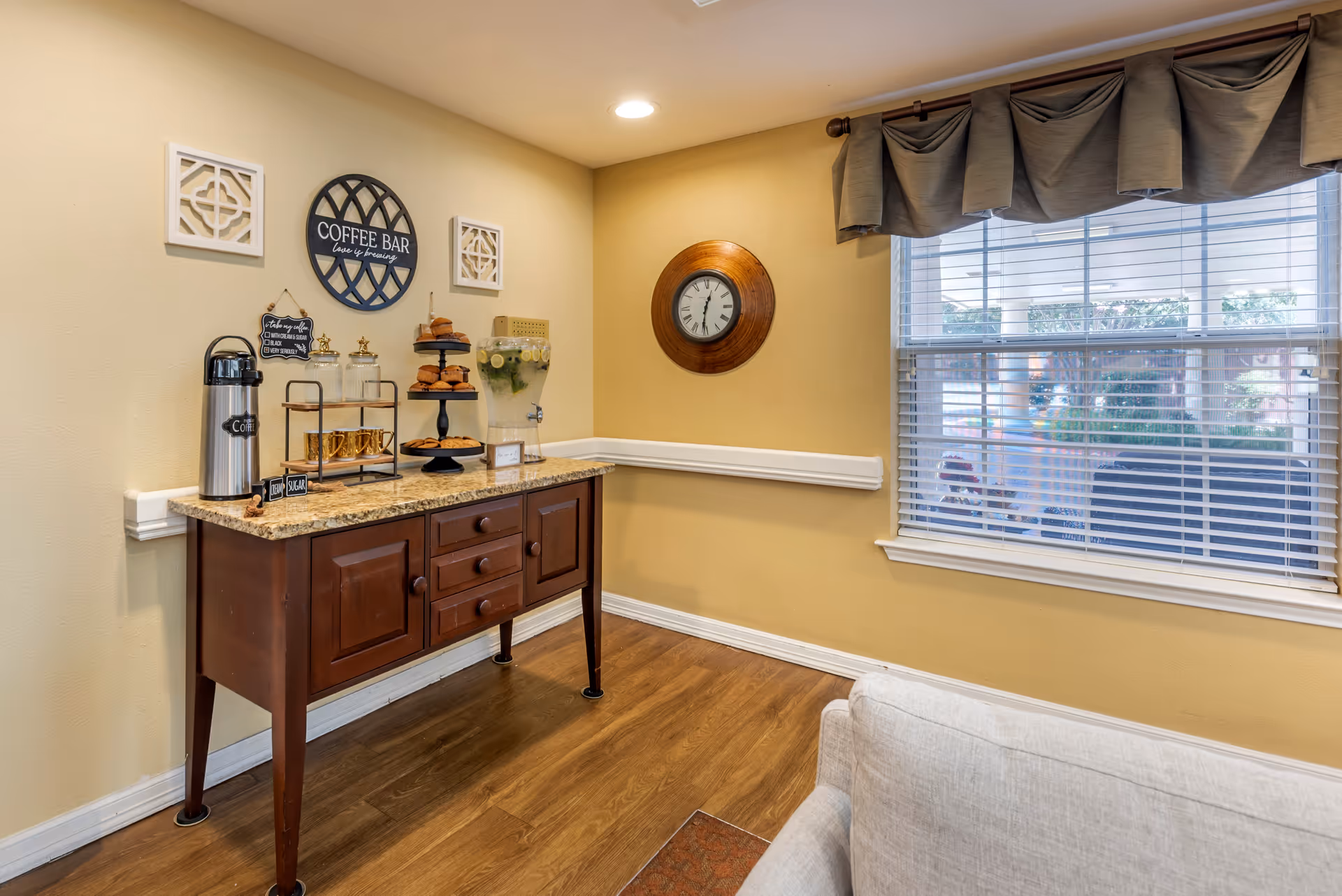 A cozy corner of a room featuring a wooden coffee bar table with coffee supplies, pastries, and a glass dispenser with lemon water. Above the table are decorative wall hangings including a round sign that says 'COFFEE BAR love is brewing'. A round wooden clock is mounted on the adjacent wall. A window with blinds and a valance lets in natural light, and part of a light-colored sofa is visible in the foreground.