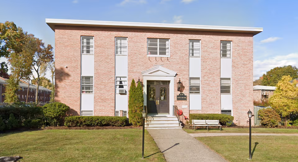 Front exterior view of a two-story brick building with multiple windows, a central entrance with double doors, a small set of stairs, and a pathway leading to the entrance. The building is surrounded by grass, bushes, trees, and a bench near the entrance.