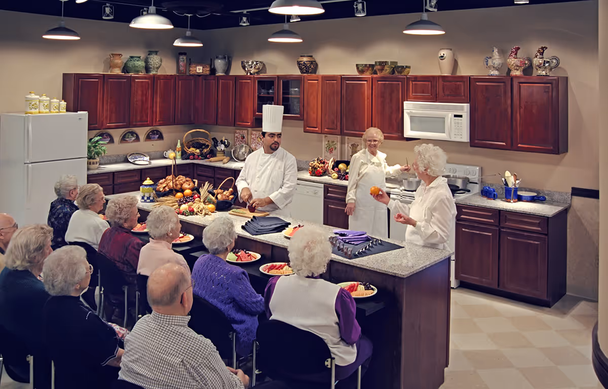 A group of elderly people seated and watching a cooking demonstration in a kitchen. A chef in a white uniform and hat is preparing food on the counter, while two elderly women stand nearby, one holding an orange. The kitchen has dark wooden cabinets, a white refrigerator, a microwave, and various decorative items on the countertops and shelves.
