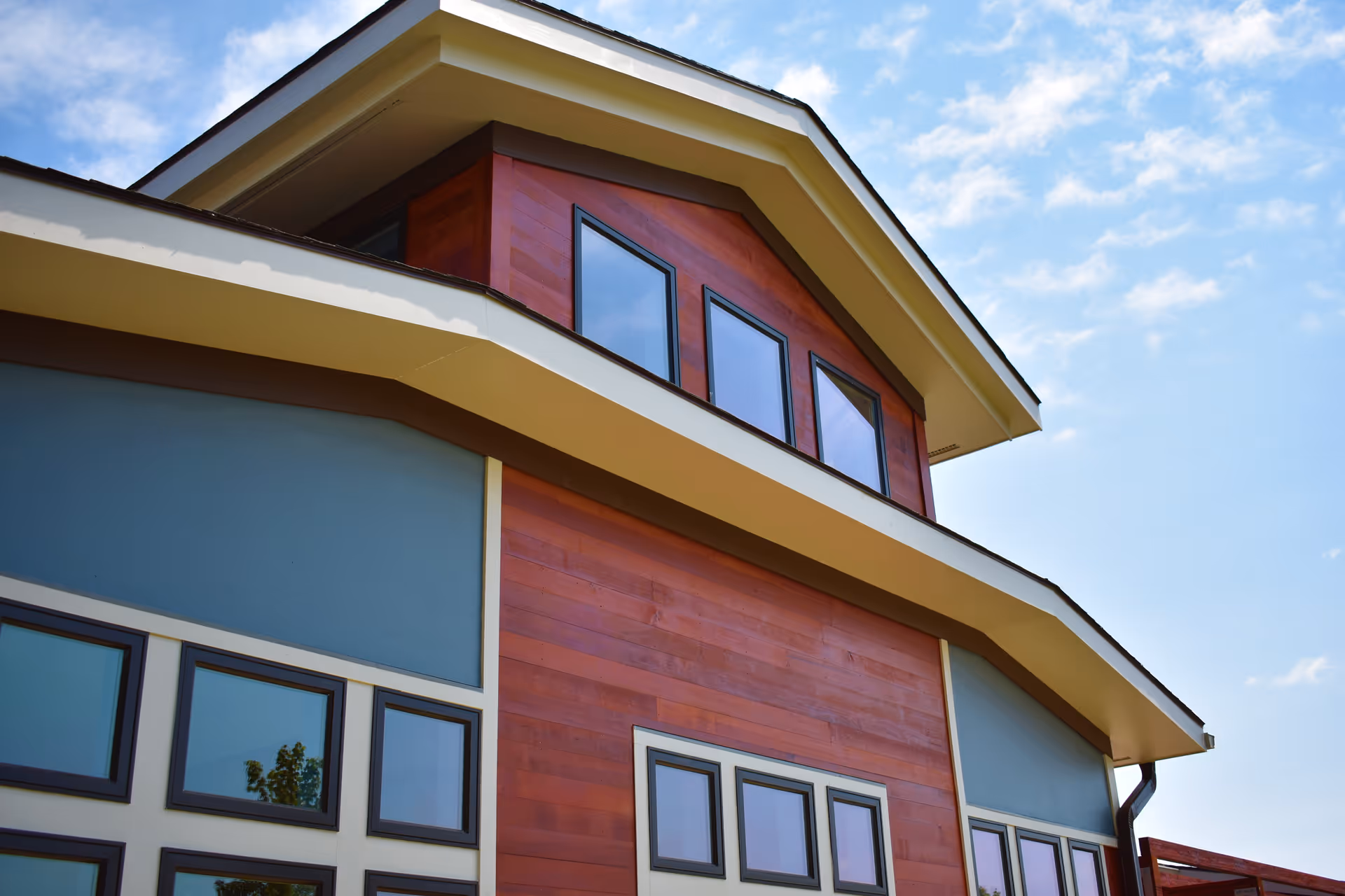 Upward view of a modern building exterior with a combination of red wood paneling and blue-gray walls, featuring multiple rectangular windows and a clear blue sky with some clouds in the background.