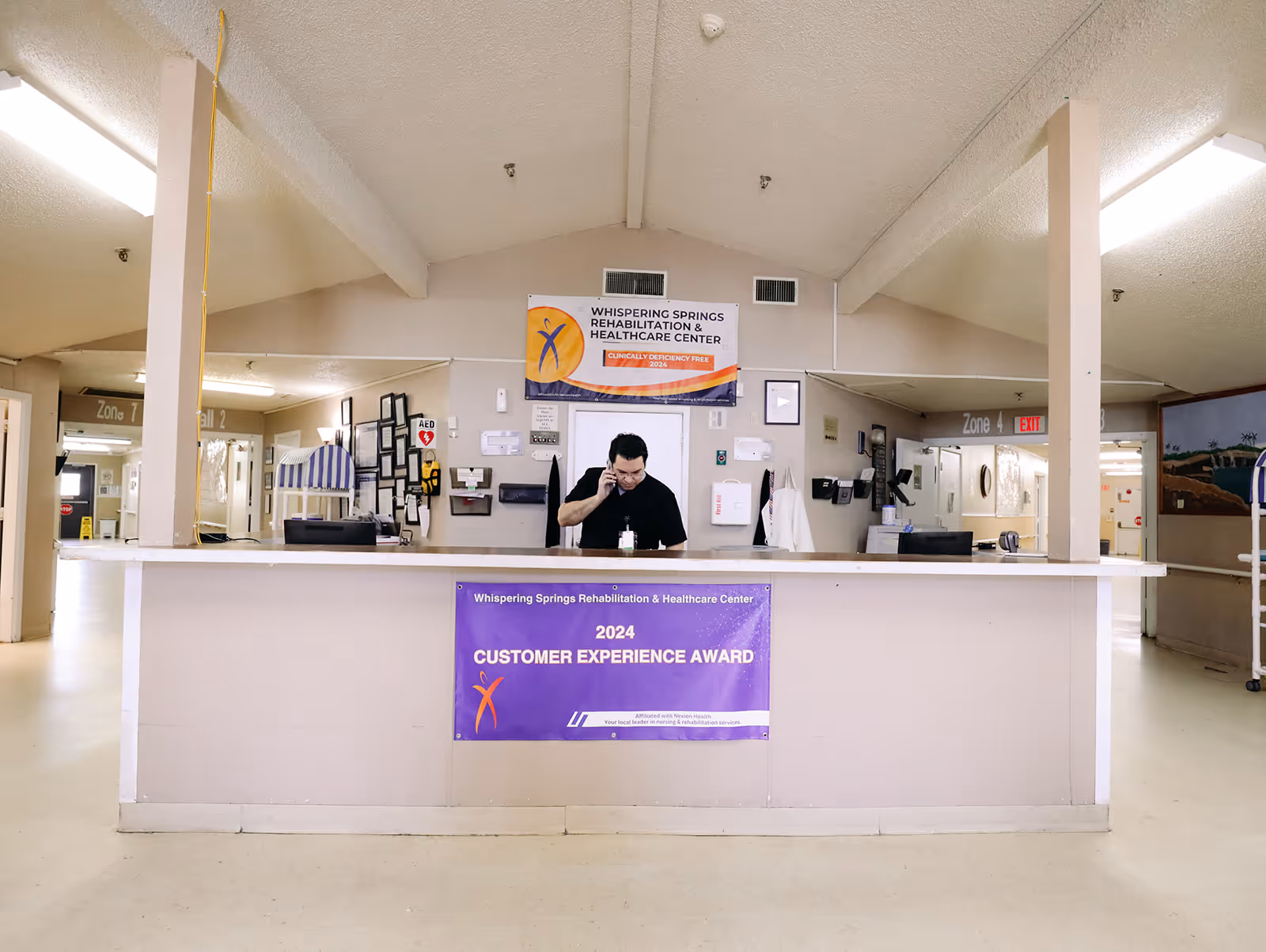 Reception desk area inside Whispering Springs Rehabilitation and Healthcare Center with a staff member standing behind the desk talking on the phone. There are signs and awards displayed on the wall and desk, and hallways leading to different zones are visible in the background.