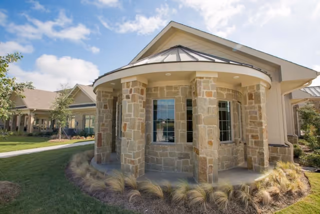 Exterior view of a building with stone pillars and a rounded porch area, surrounded by grass and landscaping under a partly cloudy sky.