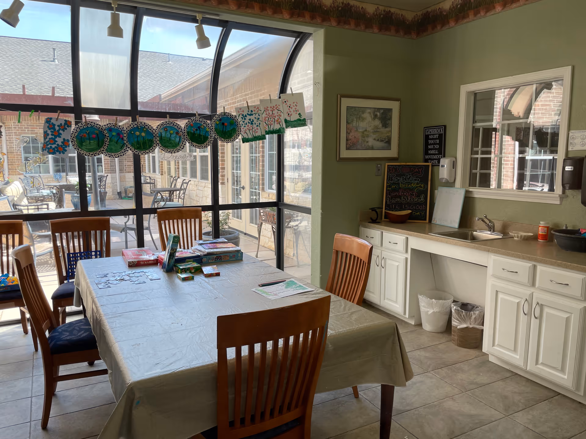 A bright room with a large table covered with a plastic tablecloth and surrounded by wooden chairs. On the table are various board games and puzzle pieces. The room has large windows with a view of an outdoor patio area with tables and chairs. Artwork and crafts are displayed hanging near the windows. There is a countertop with a sink, cabinets, a chalkboard with colorful writing, and a soap dispenser on the wall.