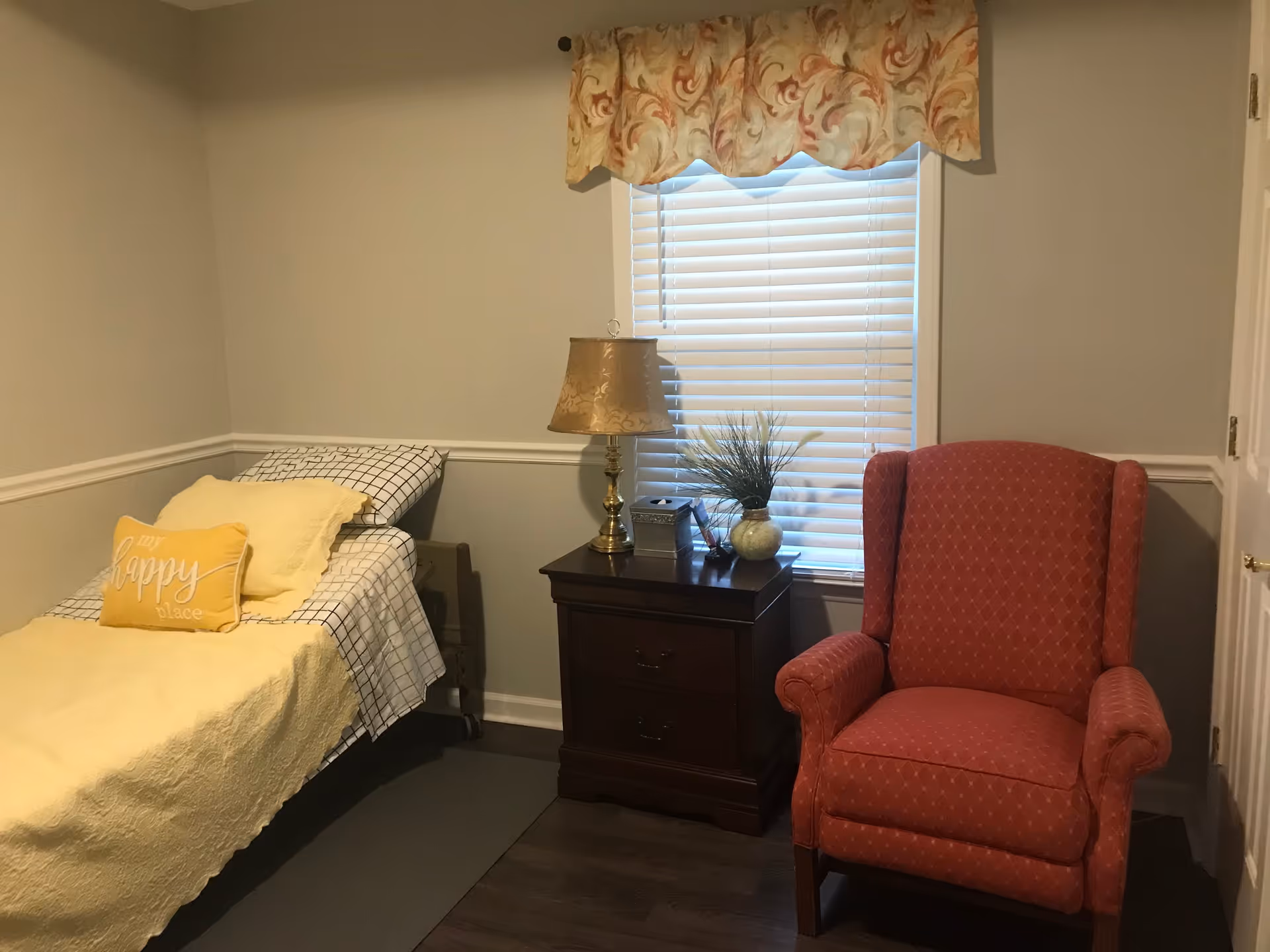 Small furnished bedroom with a single bed, nightstand with a lamp and decor, and a red upholstered armchair by a window.