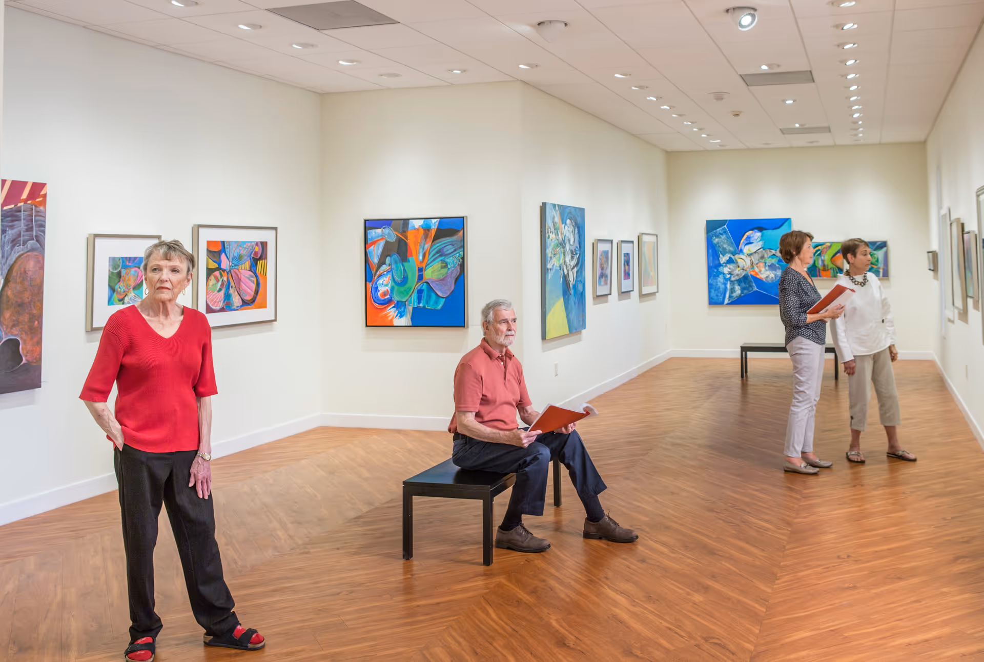 An art gallery room with wooden flooring and white walls displaying colorful abstract paintings. Four elderly individuals are present; one woman in a red top and black pants stands near the left wall, a man in a red shirt and dark pants sits on a bench holding a booklet, and two women on the right side are looking at the artwork while holding booklets.