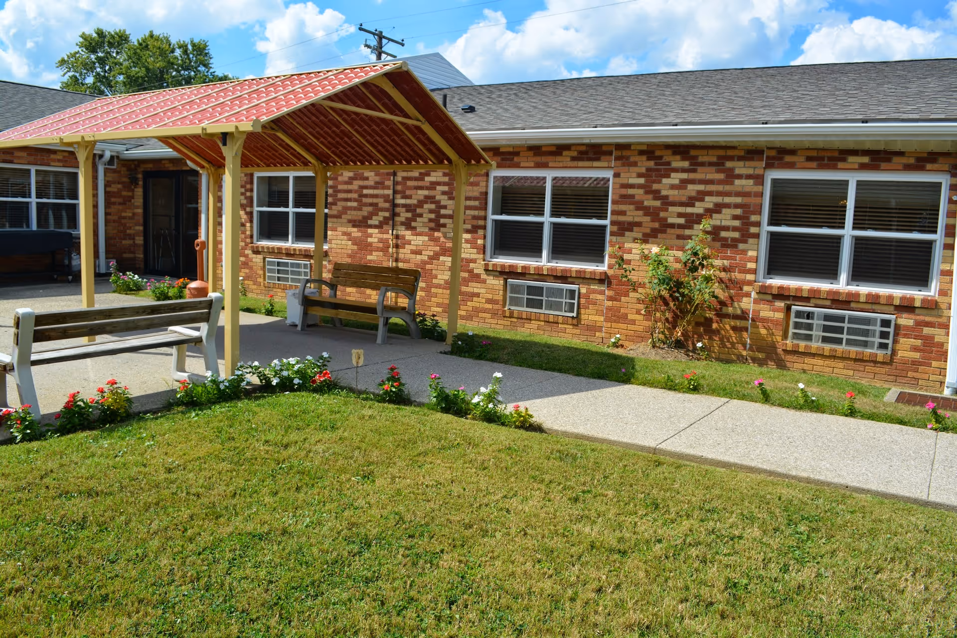 Outdoor area of a senior living facility with a small covered seating area featuring two wooden benches. The building has a brick exterior with multiple windows and air conditioning units below them. There is a concrete pathway and a well-maintained lawn with flower beds along the edges.