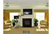 A bright lounge area with a fireplace in the center, a flat-screen TV mounted above it, two armchairs on either side, large windows with yellow curtains, and two potted plants flanking the fireplace.