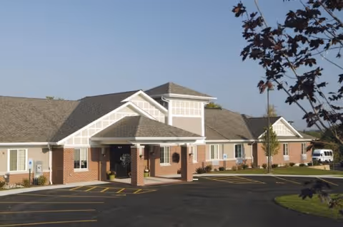 Exterior view of a single-story brick building with a covered entrance, multiple windows, and a parking lot in front. There are small trees and landscaping around the building under a clear blue sky.