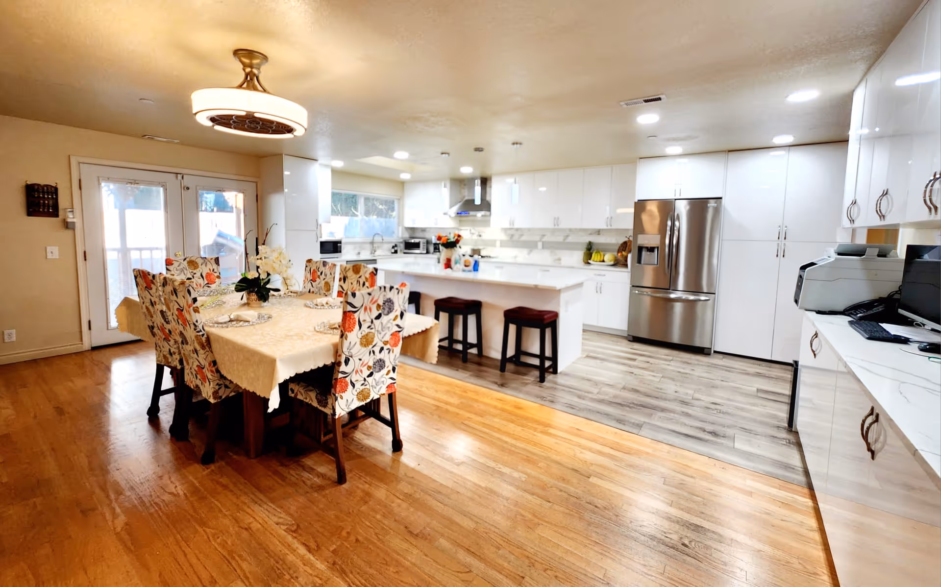 A bright and spacious kitchen and dining area with wooden floors and white cabinetry. The dining table is covered with a beige tablecloth and surrounded by six floral-patterned chairs. The kitchen features a large island with two stools, stainless steel refrigerator, and modern appliances. French doors with glass panels lead outside, allowing natural light to fill the room.