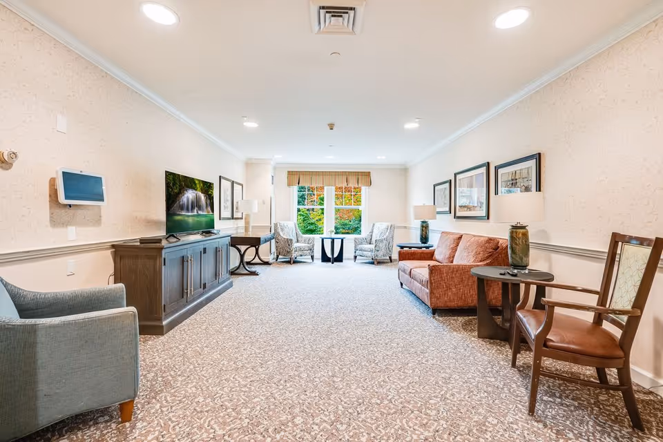 A bright and spacious living room area in a senior living facility with a patterned carpet, beige walls, and recessed ceiling lights. The room features a large flat-screen TV on a wooden cabinet, a blue armchair, two patterned armchairs near a window with a view of greenery, a rust-colored sofa, wooden side tables with lamps, and framed artwork on the walls.