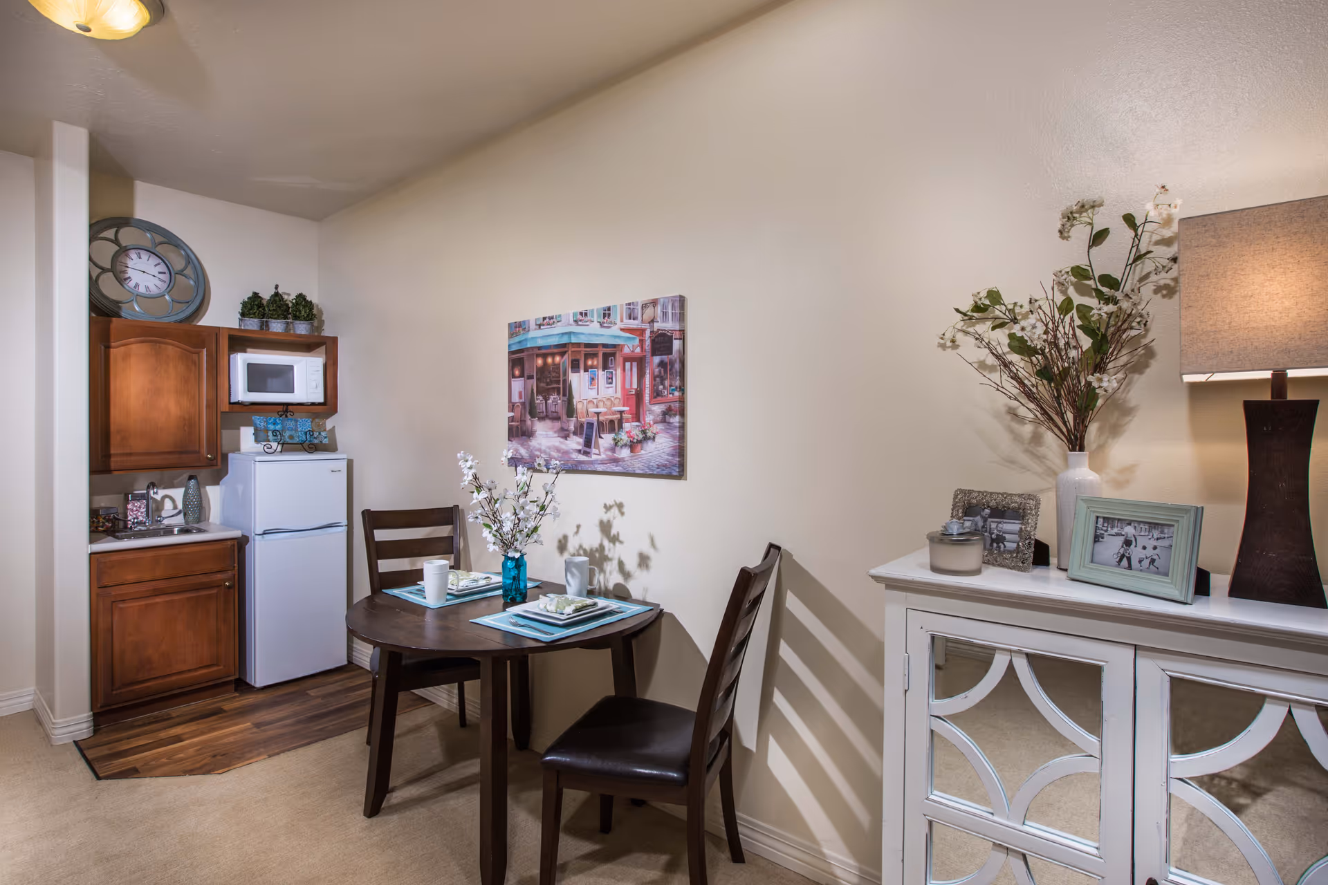 Small dining area with a round wooden table set for two with placemats, plates, and cups. Behind the table is a kitchenette with wooden cabinets, a white microwave, and a white refrigerator. On the right side, there is a white cabinet with mirrored doors, decorated with a vase of flowers, framed photos, a candle, and a lamp. A painting of a street cafe hangs on the wall above the dining table.