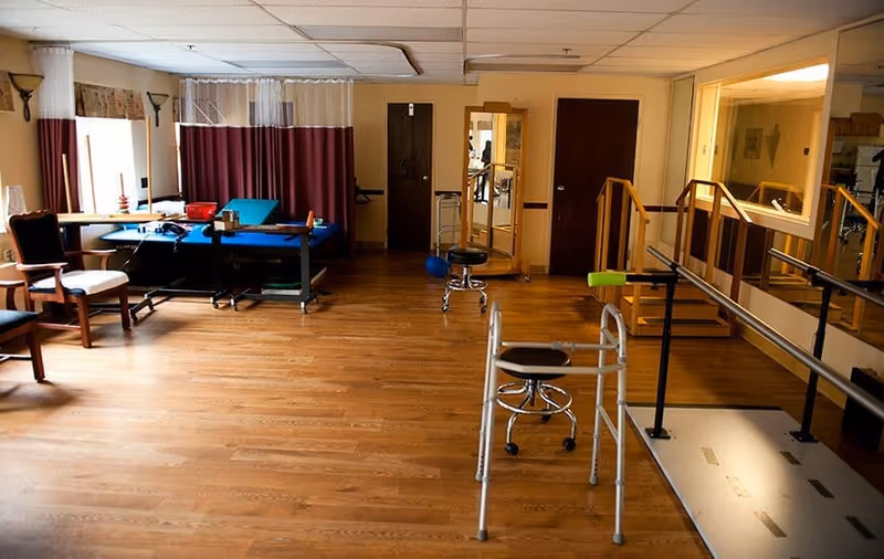 A rehabilitation room with wooden flooring featuring parallel bars for walking exercises, a walker, stools, chairs, a treatment table, and a mirror. The room has windows with curtains and a partition curtain in the background.
