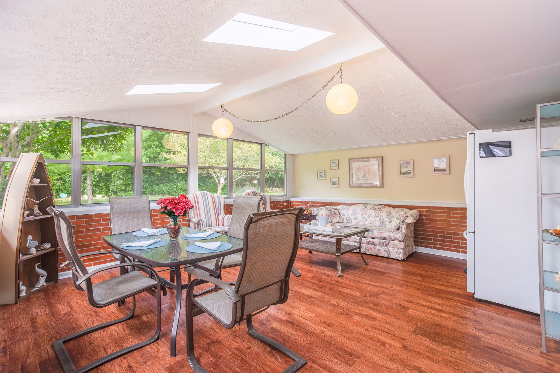 A bright and cozy living room area with large windows showing green trees outside. The room features a glass dining table with four chairs, a vase with red flowers on the table, two striped armchairs, a floral-patterned sofa, a glass coffee table, and a white refrigerator. The ceiling has two round hanging light fixtures and skylights, and the floor is wooden.