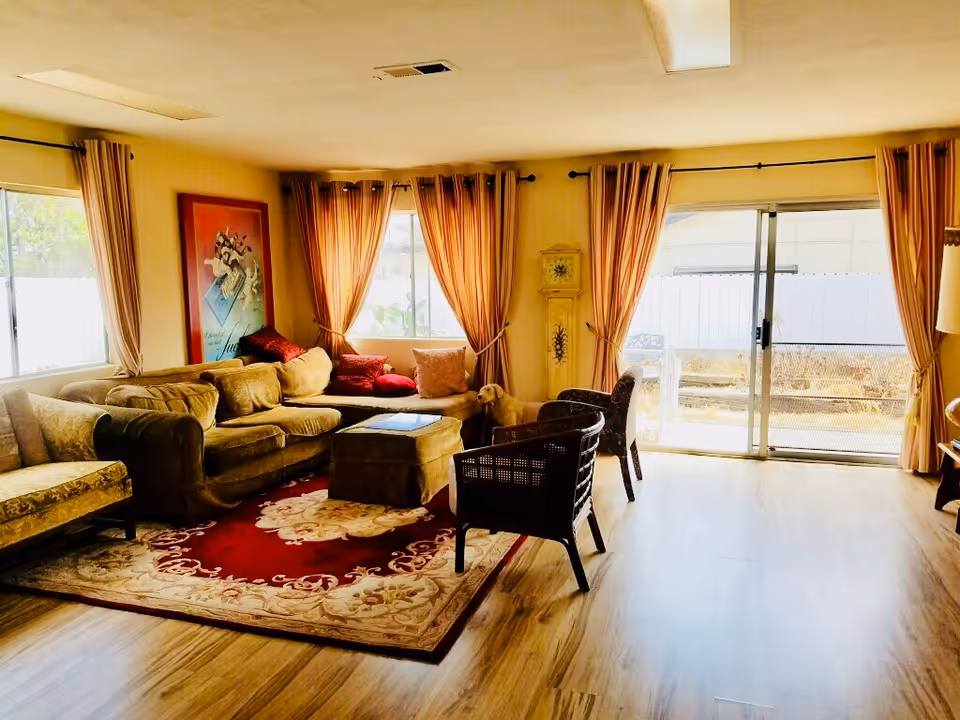 Cozy senior living living room with beige sofas, chairs, rug, and sliding glass door.