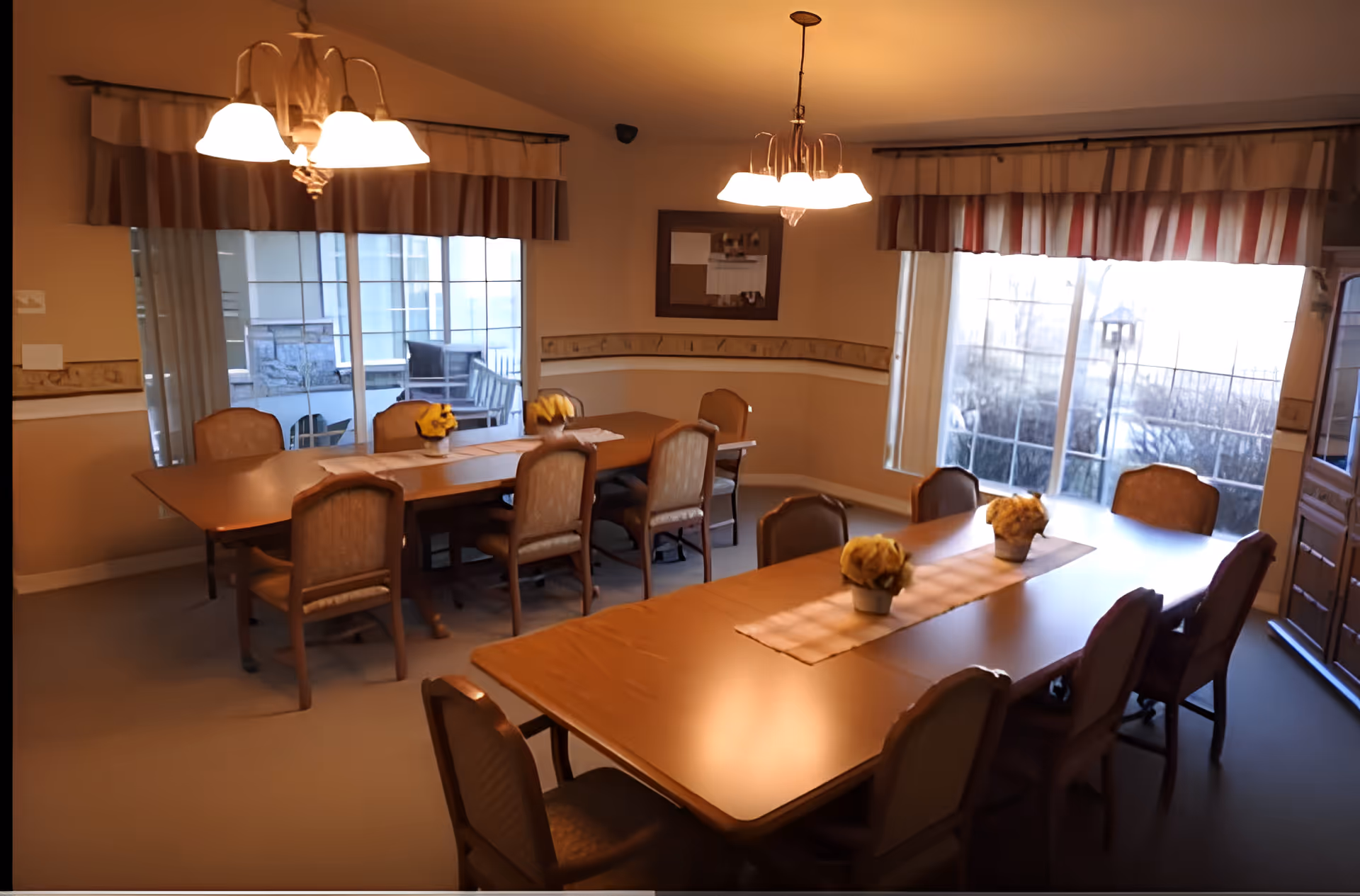 A dining room with two wooden tables, each surrounded by chairs. The tables have beige runners and small flower arrangements. Large windows with striped valances let in natural light, and two ceiling light fixtures illuminate the room. There is a bulletin board on the wall and a wooden cabinet in the corner.