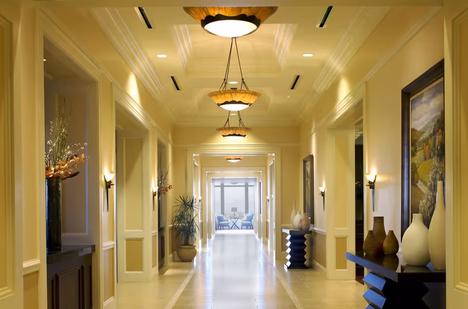 A well-lit, elegant hallway with cream-colored walls and ceiling, decorated with modern hanging light fixtures and wall sconces. The hallway features decorative vases and framed artwork on the walls, with a seating area visible at the far end near large windows.