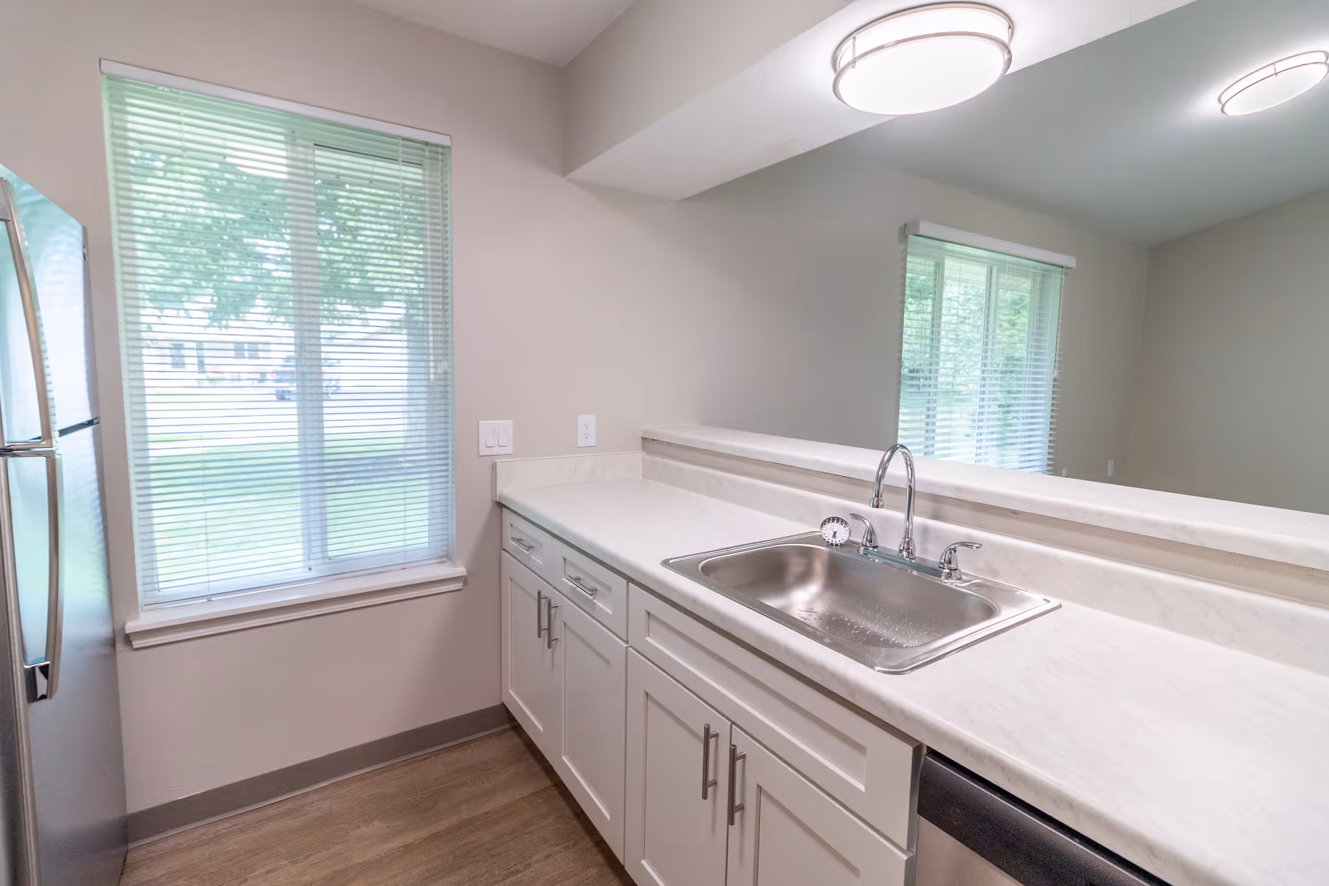 A clean and modern kitchen area featuring white cabinets with silver handles, a stainless steel sink with a chrome faucet, and a large window with blinds letting in natural light. The countertop is light-colored, and a stainless steel refrigerator is partially visible on the left side. The room has light-colored walls and wood-style flooring.