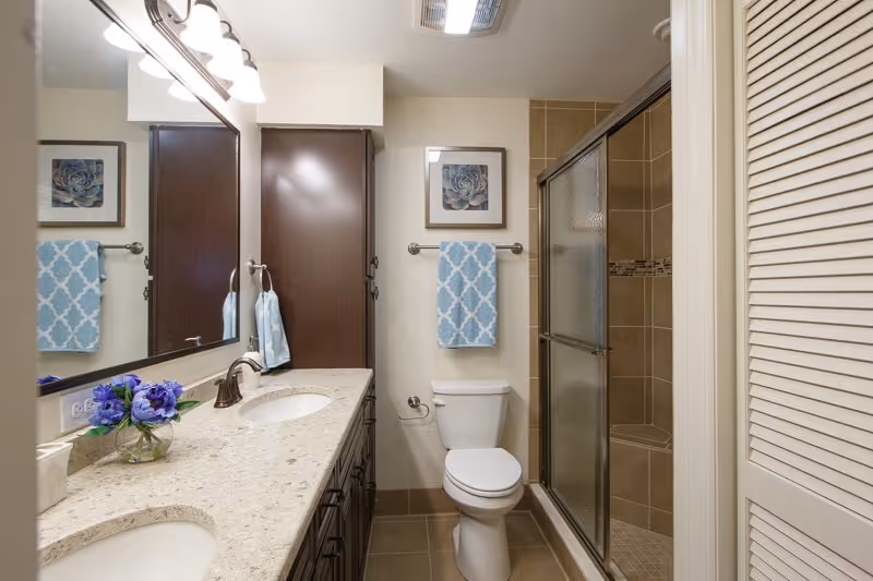 A clean and modern bathroom featuring a double sink vanity with a granite countertop, a large mirror, a toilet, and a glass-enclosed shower with tiled walls. There is a blue patterned towel hanging on a towel rack above the toilet and a small vase with purple flowers on the countertop. A framed floral artwork is hung on the wall above the towel rack.