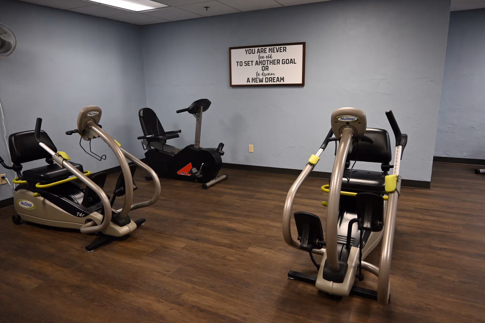 Exercise room with recumbent cardio machines on a wood floor and a motivational sign on the wall.