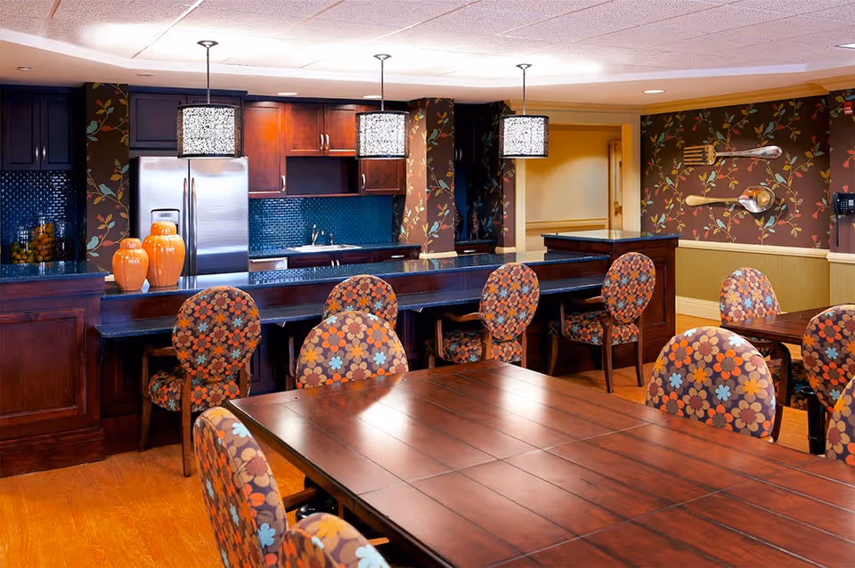 Bright dining area with floral-upholstered chairs around wooden tables and a serving bar/kitchen with pendant lights.