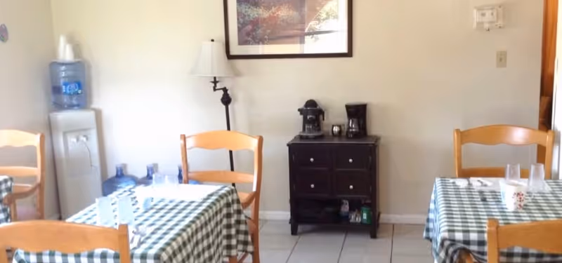 A small dining area with two tables covered in green and white checkered tablecloths, each set with plastic cups and utensils. Four wooden chairs are around the tables. Against the wall is a water dispenser with a large water bottle on top and an extra bottle on the floor. A small dark wooden cabinet holds a coffee maker and other small items. A framed picture hangs above the cabinet, and a floor lamp is positioned next to it.