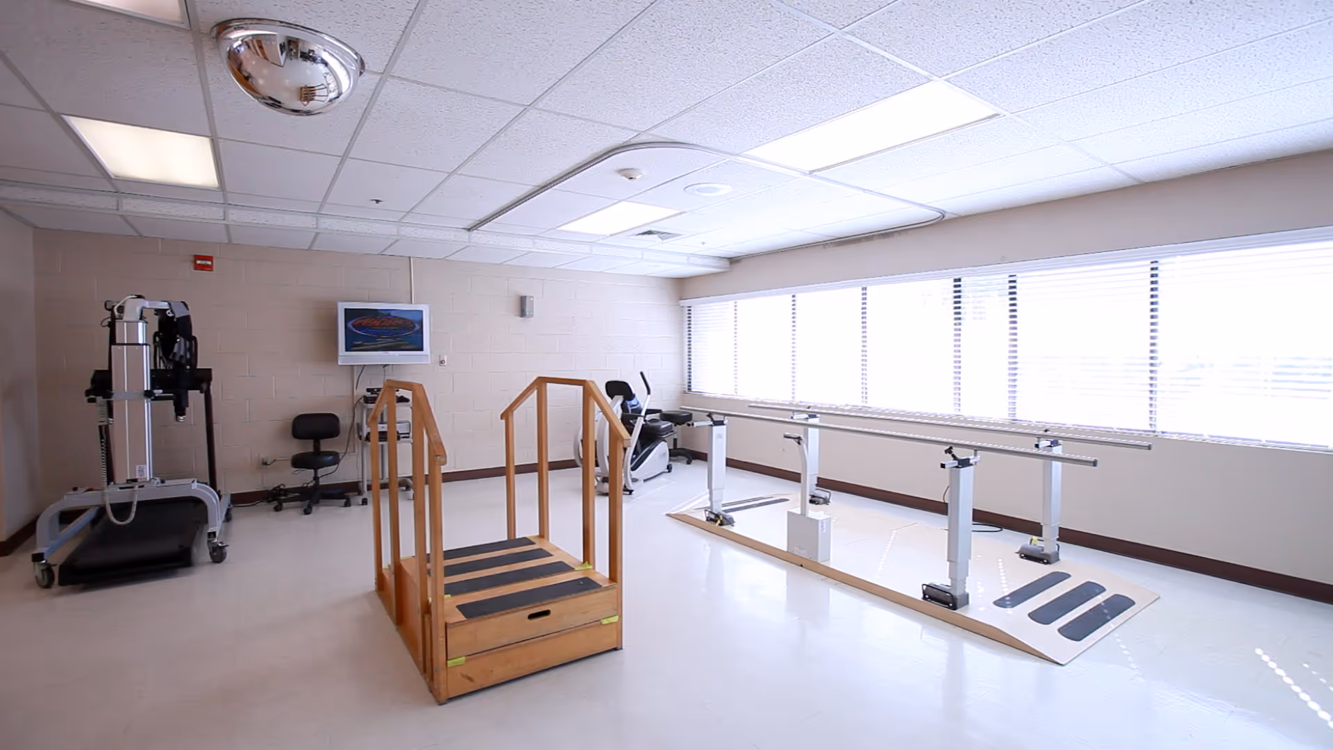 A bright physical therapy room with large windows covered by blinds. The room contains a wooden step platform with handrails, parallel bars for walking exercises, a stationary exercise bike, and a piece of exercise equipment with a chair. The floor is clean and white, and the ceiling has fluorescent lights.