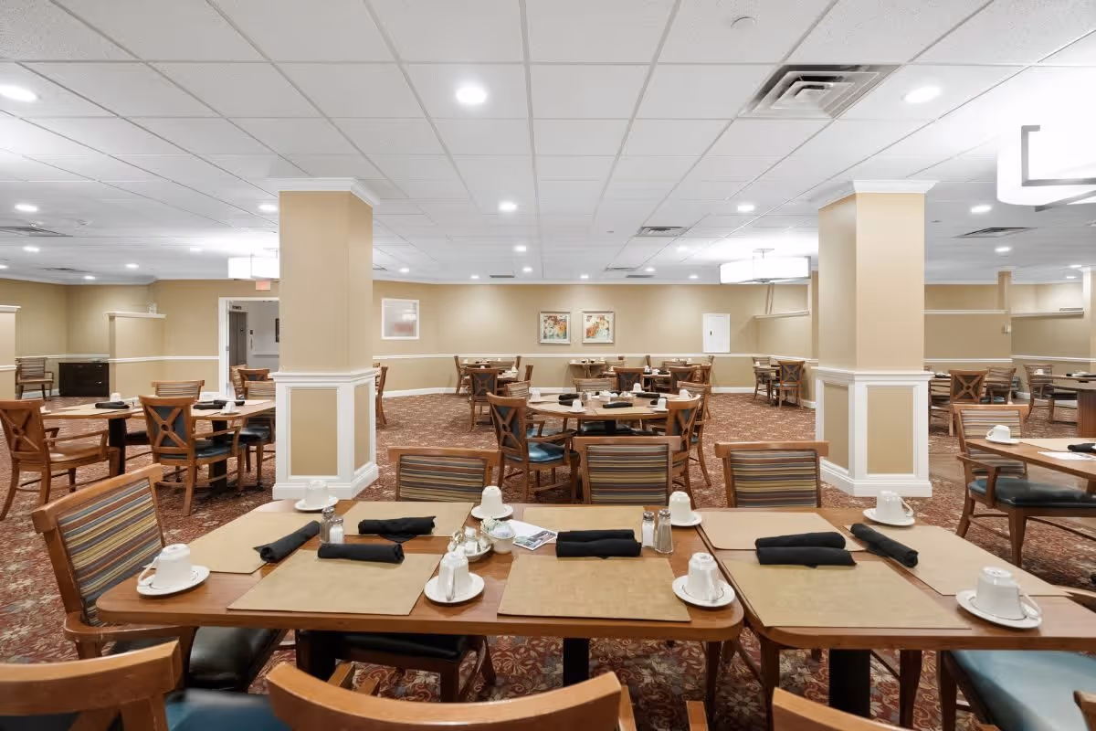 A spacious dining room with multiple wooden tables and chairs arranged neatly. Each table is set with placemats, cups, saucers, and black napkins. The room has beige walls, white ceiling tiles with recessed lighting, and carpeted floors with a patterned design. There are two large columns in the center of the room and framed artwork on the far wall.