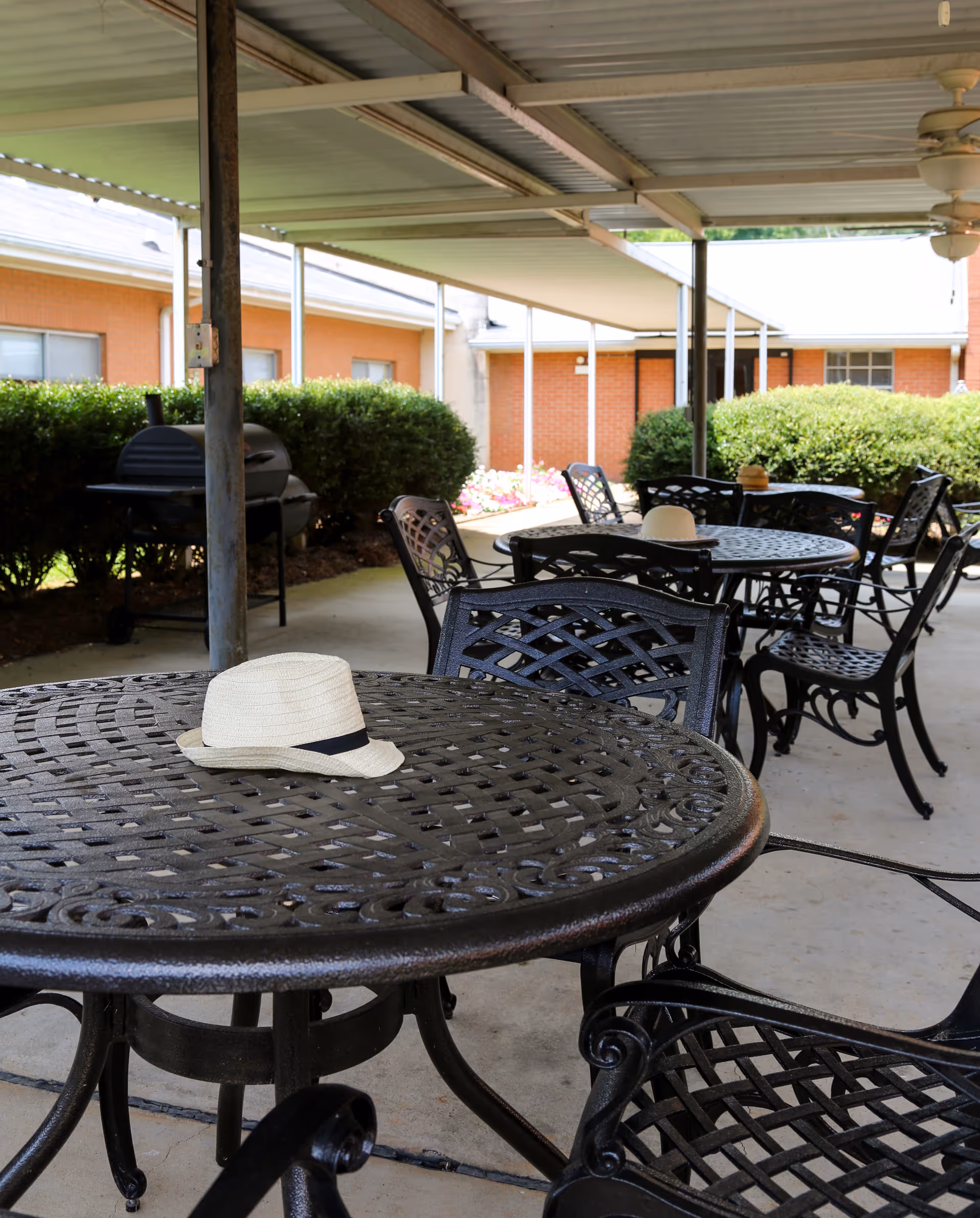 Covered outdoor patio with ornate metal tables and chairs, a grill, and straw hats on the tables in front of a brick building.