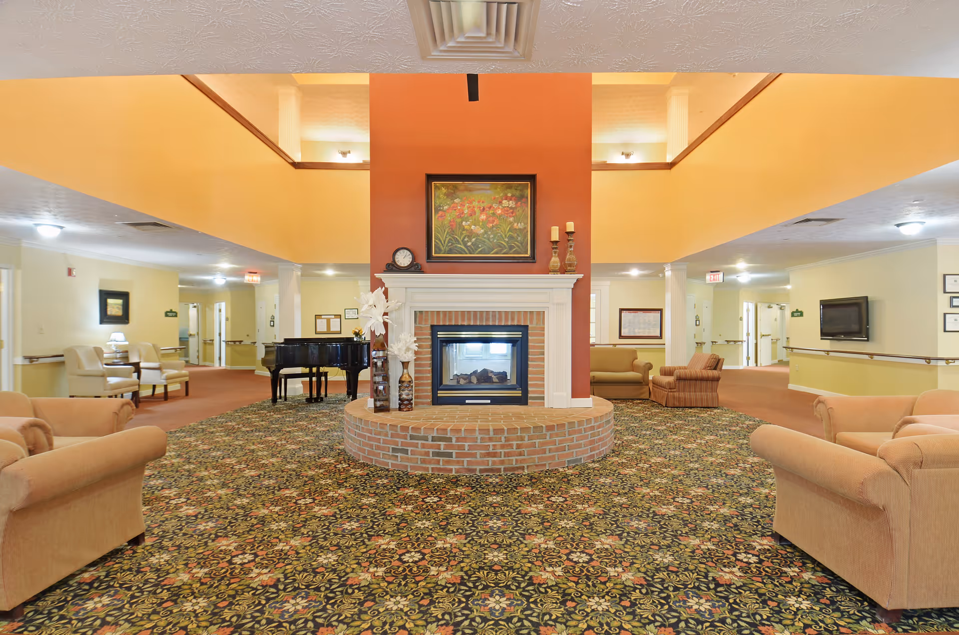 A spacious senior living facility common area with a central brick fireplace surrounded by beige armchairs and sofas. The room features a floral patterned carpet, a grand piano, and warm yellow walls with white trim. There is a painting above the fireplace and decorative vases on the hearth. The ceiling has recessed lighting and a second-floor balcony overlooking the area.