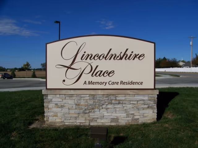 A large outdoor sign for Lincolnshire Place, a memory care residence, mounted on a stone base with a clear blue sky and some trees and a car in the background.
