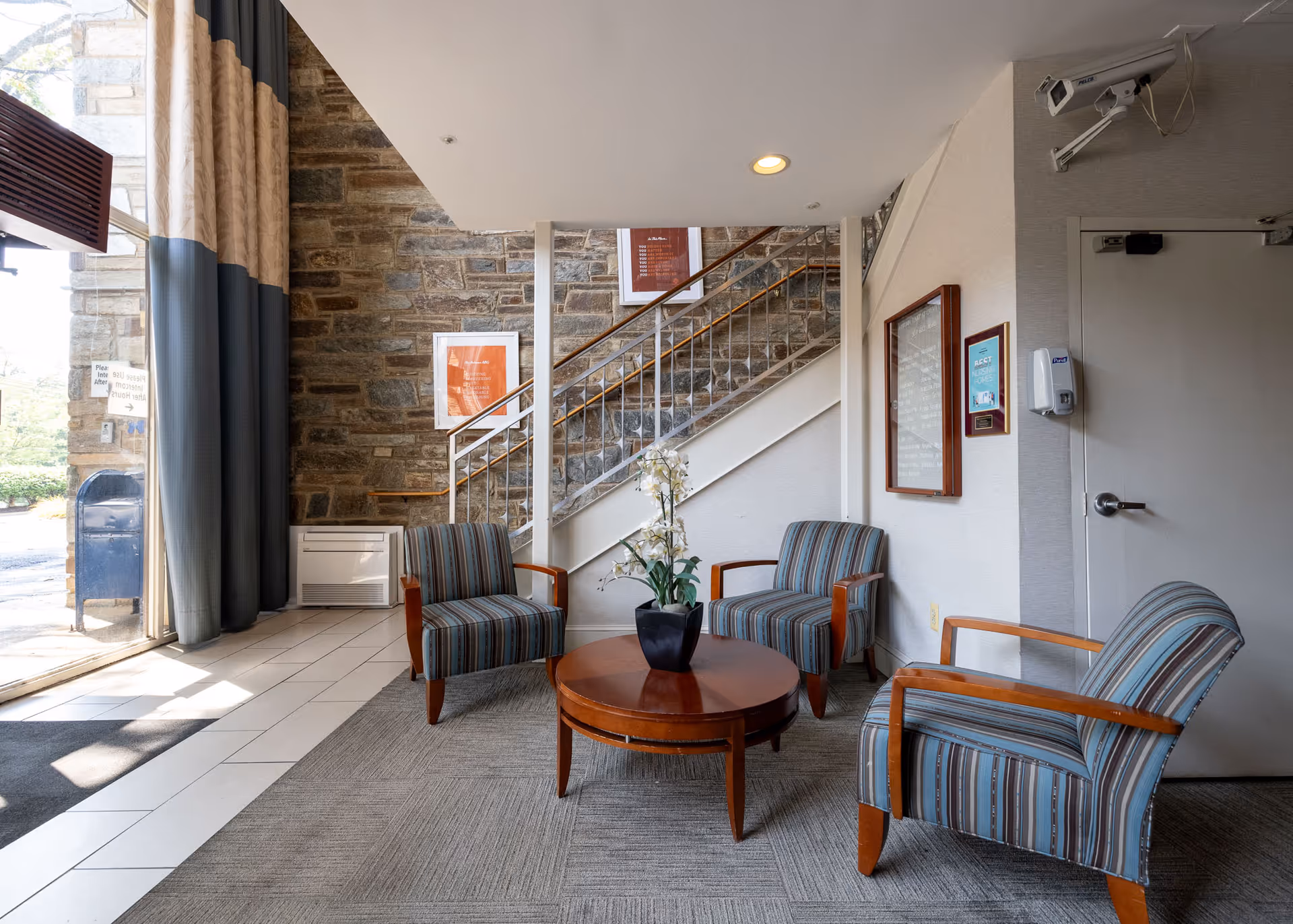 A small seating area inside a facility with three striped armchairs arranged around a round wooden coffee table with a potted orchid plant. Behind the seating area is a stone wall with framed artwork and a staircase with a metal railing. To the left, large windows with curtains allow natural light to enter. On the right wall, there is a door, a hand sanitizer dispenser, and framed notices.