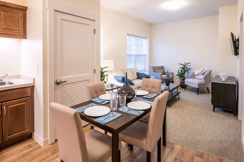 A cozy interior space featuring a dining area with a dark wooden table set for four with plates, glasses, and blue napkins. Adjacent to the dining area is a living room with a beige sofa, two armchairs, a coffee table with decorative vases, a potted plant, and a wall-mounted TV. The room has light-colored walls, a window with blinds, and wood and carpet flooring.