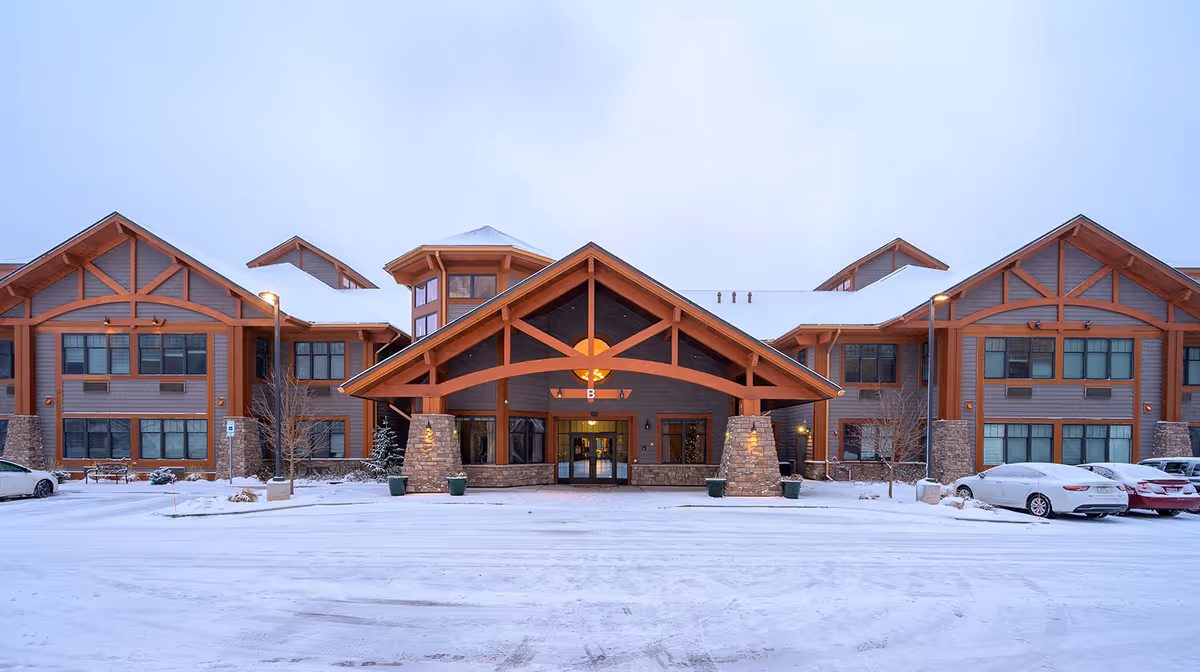 Front exterior view of Creekside Assisted Living LLC building in winter with snow covering the ground and parked cars on either side. The building features a large wooden entrance with stone pillars and multiple windows.